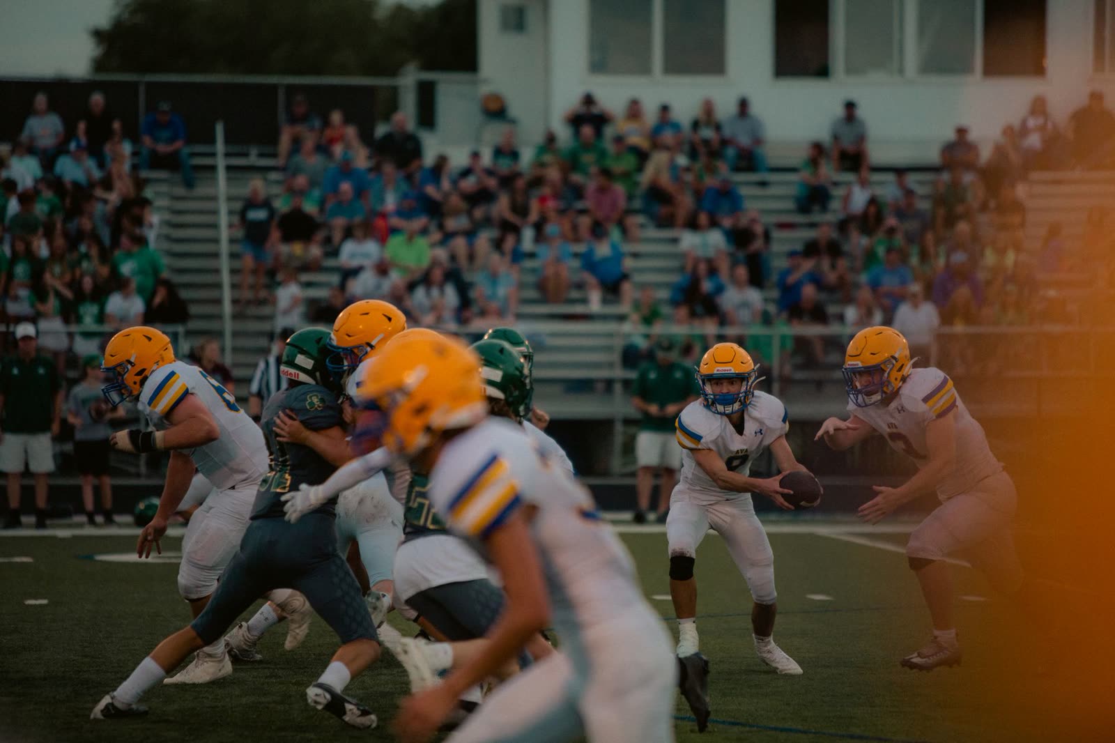 fans in short sleeves in the stands at a high school football game at night / spectators with umbrellas at a football stadium on a humid evening / high school football stadium lights glowing at dusk