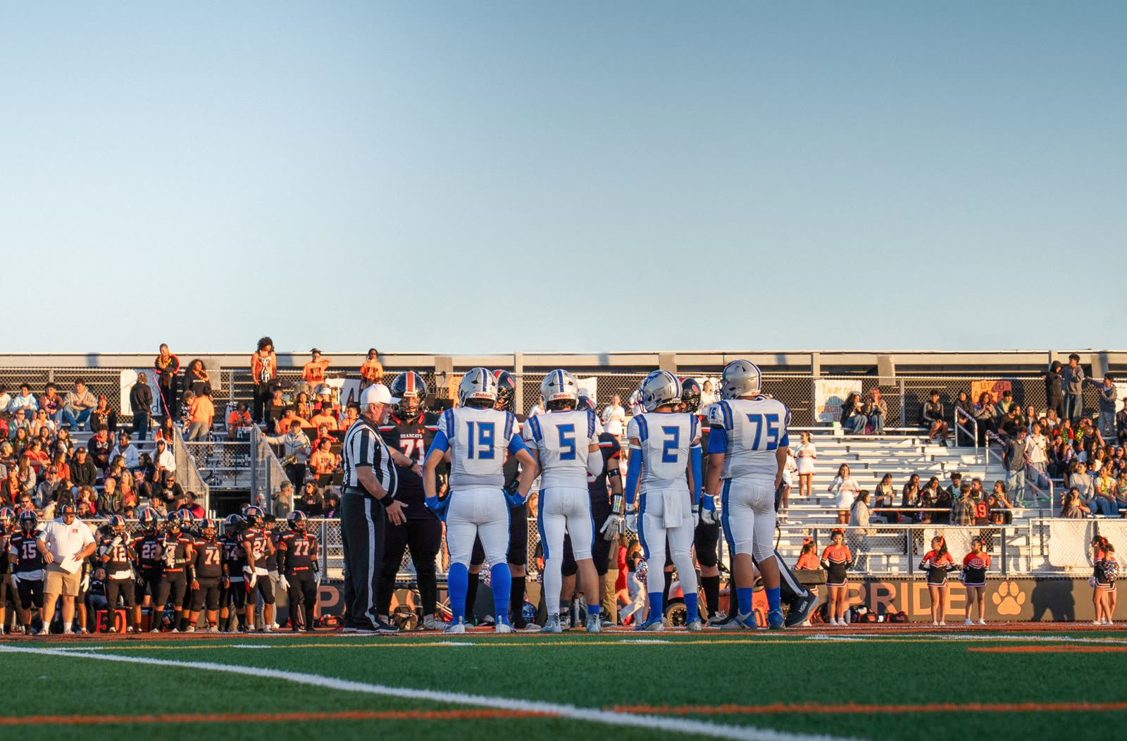 fans in hoodies in the bleachers at a high school football game at night / high school football stadium lights against a clear evening sky / high school marching band in the stands on a cool autumn night