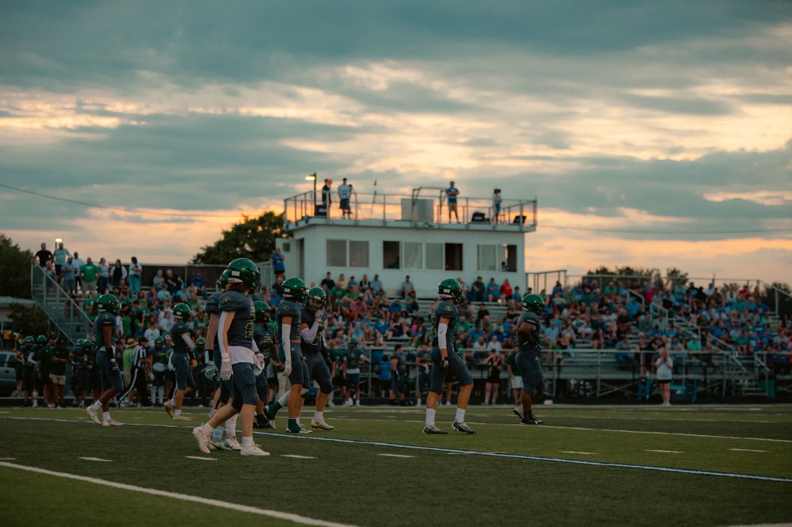 fans in hoodies in the stands at a high school football game at night / students cheering under stadium lights on a clear fall evening / high school football stadium lights against a clear night sky